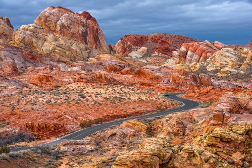 Winding road through colorful rock formations under a cloudy sky in a desert landscape.