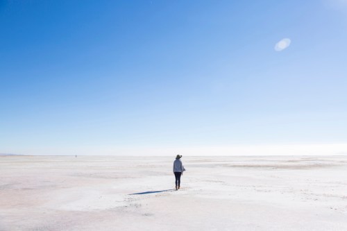 A person walks alone across a vast, empty salt flat under a clear blue sky.