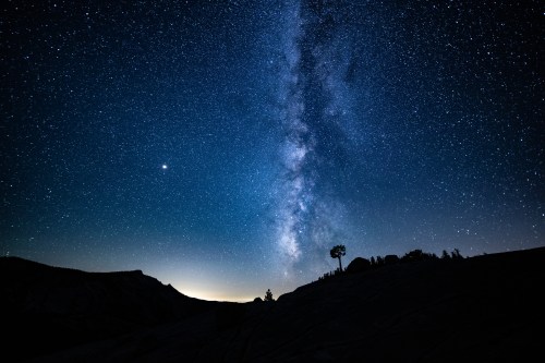 A starry night sky with the Milky Way stretching across, silhouetted trees and mountains in the foreground.