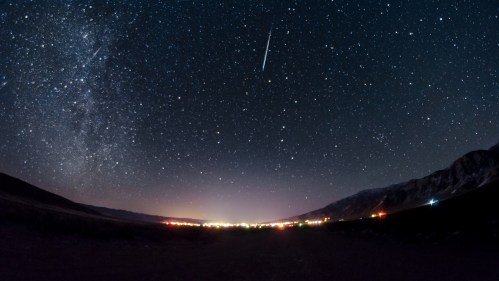 A starry night sky with a bright meteor streaking across, illuminating a distant city below.