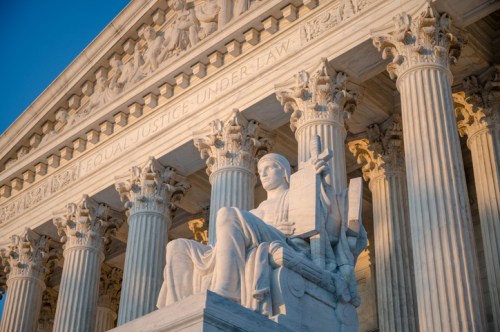 A close-up of the Supreme Court building, featuring a statue of Lady Justice and ornate columns under a clear sky.