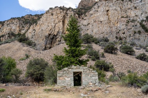 A small stone structure stands in a rocky landscape, surrounded by trees and steep cliffs in the background.
