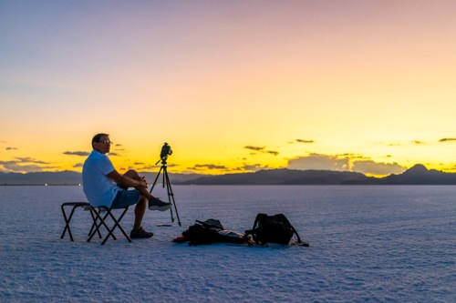 A photographer sits on a chair on a salt flat, capturing a vibrant sunset with mountains in the background.
