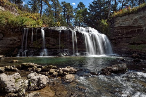 A serene waterfall cascades over rocks into a clear pool, surrounded by lush greenery and trees under a blue sky.