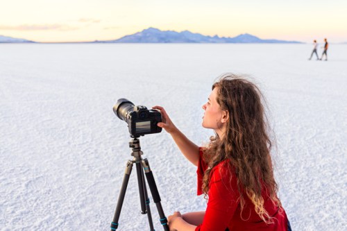 A woman in a red dress sets up a camera on a tripod in a vast, white salt flat with mountains in the background.
