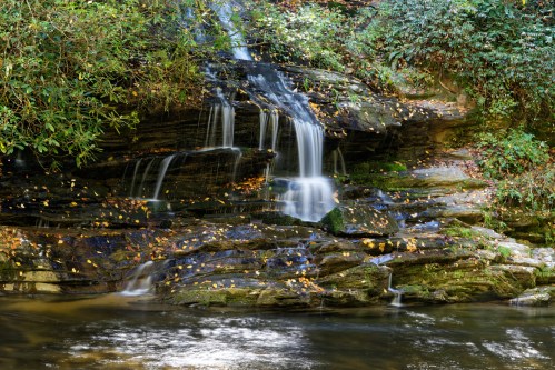 A serene waterfall cascades over rocky ledges, surrounded by lush greenery and autumn leaves.