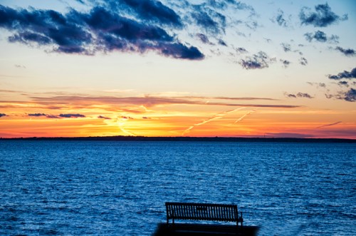 A serene sunset over a calm sea, with a lone bench in the foreground and colorful clouds in the sky.