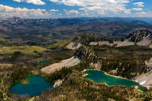 Aerial view of a mountainous landscape with vibrant green lakes and lush forests under a partly cloudy sky.