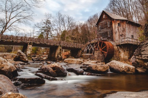A rustic wooden mill with a waterwheel beside a flowing river and a stone bridge, surrounded by trees and rocky terrain.