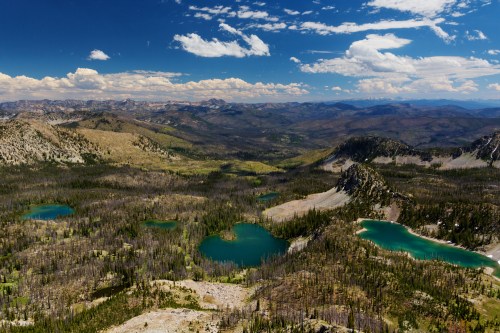 A panoramic view of mountains and vibrant blue lakes surrounded by lush greenery and scattered clouds in the sky.