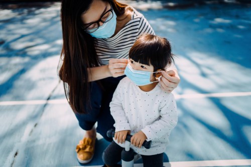 A woman helps a young child put on a face mask while both are outdoors on a blue surface.
