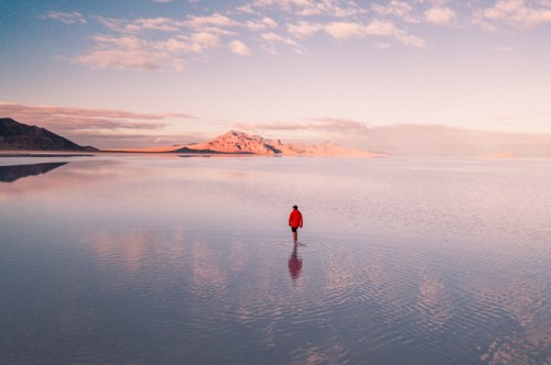 A person in a red jacket stands in shallow water, reflecting mountains and clouds in a serene landscape.