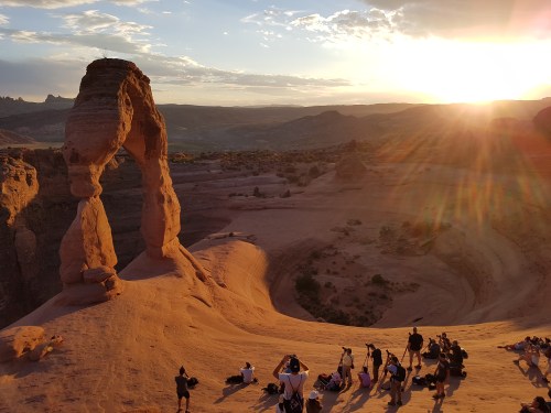 A stunning sunset over a desert landscape featuring a rock arch, with people gathered below enjoying the view.