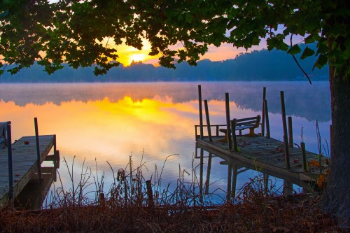 A serene lake at sunrise, with mist rising and a wooden dock framed by trees. Soft colors reflect on the water.