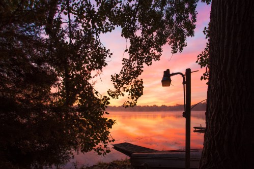 A serene lake at sunset, with colorful reflections and silhouetted trees framing a peaceful scene.