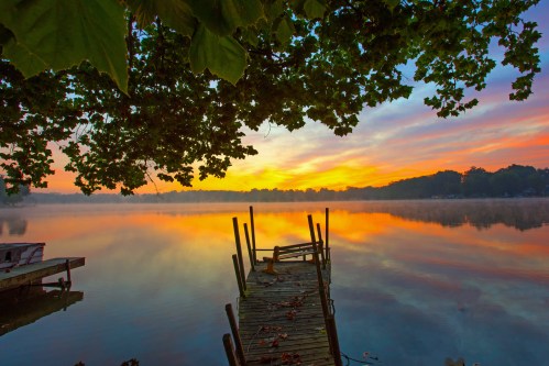 A serene lake at sunrise, with a wooden dock extending into calm waters and colorful clouds reflecting on the surface.