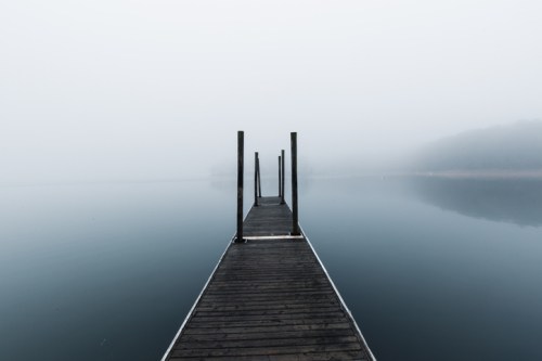 A foggy scene featuring a wooden dock extending into a calm, reflective lake.