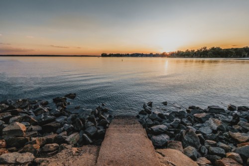 A serene lakeside view at sunset, with calm waters and a rocky shore under a colorful sky.