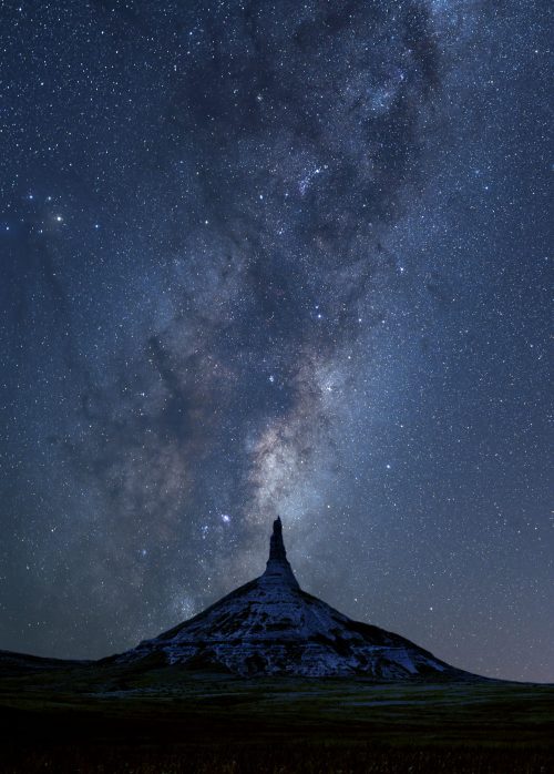 A towering rock formation under a starry night sky, with the Milky Way prominently visible above.