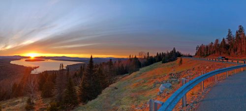 A panoramic view of a sunset over a lake, surrounded by trees and rolling hills, with a road in the foreground.