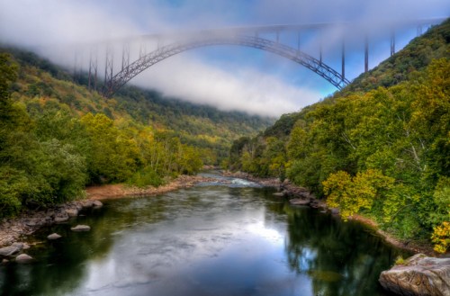 A misty river scene with a towering bridge arching over lush, green hills and trees.