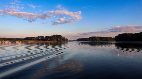 A serene lake at sunset, with gentle ripples and a clear blue sky dotted with clouds, surrounded by trees.