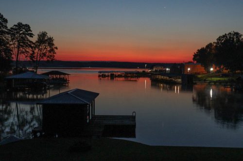 A serene lake at sunset, with colorful skies reflecting on the water and silhouettes of trees and docks.