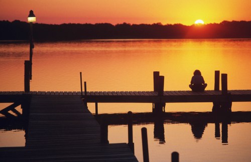 A person sits on a dock at sunset, reflecting over calm waters with vibrant orange and yellow hues in the sky.