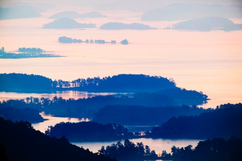 A serene landscape of misty islands on a calm lake at sunrise, with soft hues of blue and orange in the sky.