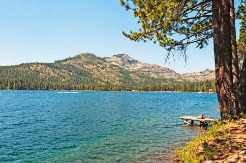 A serene lake surrounded by mountains and pine trees, with a small dock extending into the water on a sunny day.