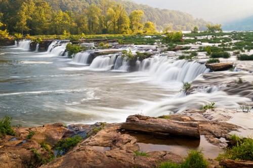 A serene waterfall cascades over rocks, surrounded by lush greenery and a tranquil river under soft sunlight.