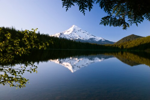 A serene lake reflects a snow-capped mountain, surrounded by lush green trees under a clear blue sky.