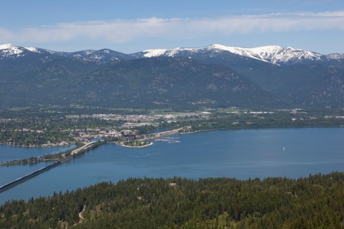 Aerial view of a town by a lake, surrounded by green forests and snow-capped mountains under a clear blue sky.
