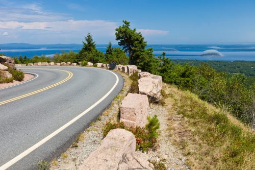 A winding road curves along a scenic overlook, with lush greenery and a distant view of water and clouds.