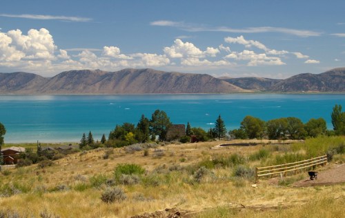 A scenic view of a turquoise lake surrounded by mountains and lush greenery under a blue sky with fluffy clouds.