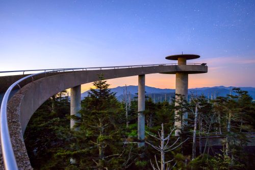 A curved observation deck above a forest, with mountains in the background and a starry sky at dusk.