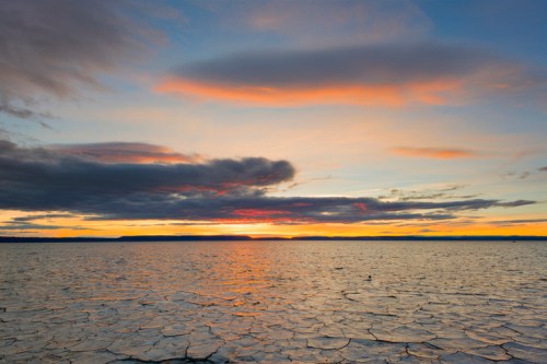 A serene sunset over a calm, icy lake with cracked ice and colorful clouds in the sky.