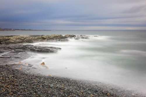 A serene coastal scene with smooth waves gently lapping against rocky shores under a cloudy sky.