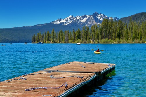 A wooden dock extends into a clear blue lake, with a kayaker paddling and mountains in the background under a clear sky.