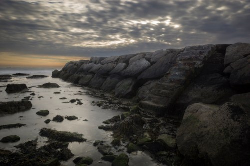 A rocky jetty extends into a calm sea under a cloudy sky, with a staircase leading down to the water's edge.
