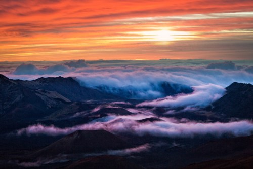 A stunning sunset over mountains, with vibrant orange skies and clouds rolling through the valleys below.