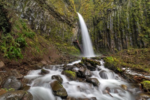 A cascading waterfall flows over moss-covered rocks, surrounded by lush greenery and steep, rocky cliffs.