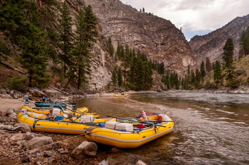 Yellow rafts are moored by a river, surrounded by rocky cliffs and tall pine trees under a cloudy sky.