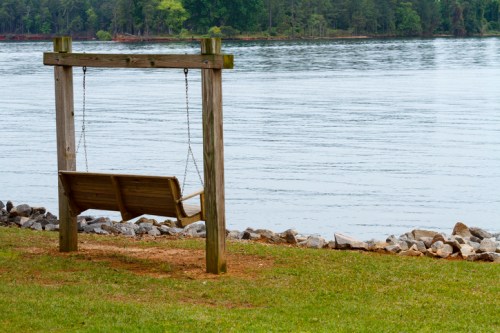 A wooden swing hangs from a frame by the water's edge, surrounded by grass and rocks, with trees in the background.
