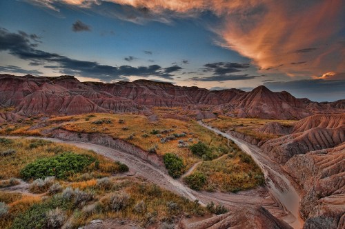 A scenic view of colorful badlands at sunset, with winding paths and patches of green vegetation among the hills.
