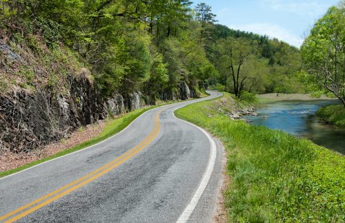 A winding road alongside a river, surrounded by lush greenery and rocky hills under a clear blue sky.