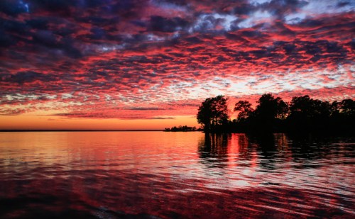 Vibrant sunset over a calm lake, with colorful clouds reflecting on the water and silhouettes of trees along the shore.