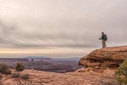 A person stands on a rocky ledge, overlooking a vast desert landscape under a cloudy sky.