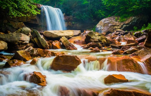 A serene waterfall cascades over rocks, surrounded by lush greenery and flowing water in a tranquil natural setting.