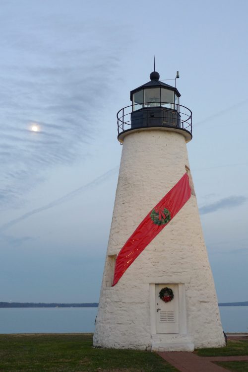 A white lighthouse adorned with a red ribbon and wreath, set against a calm sky and water.